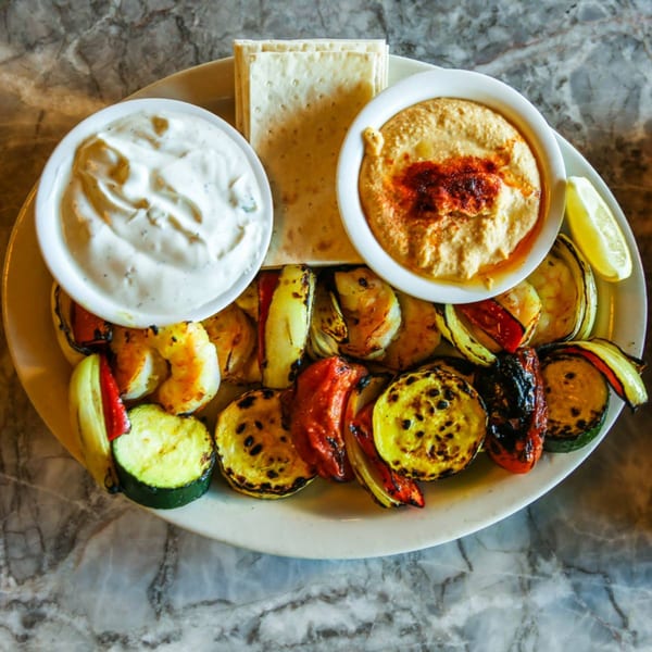 A plate of grilled vegetables, shrimp, hummus, yogurt dip, and flatbread on a marble table.