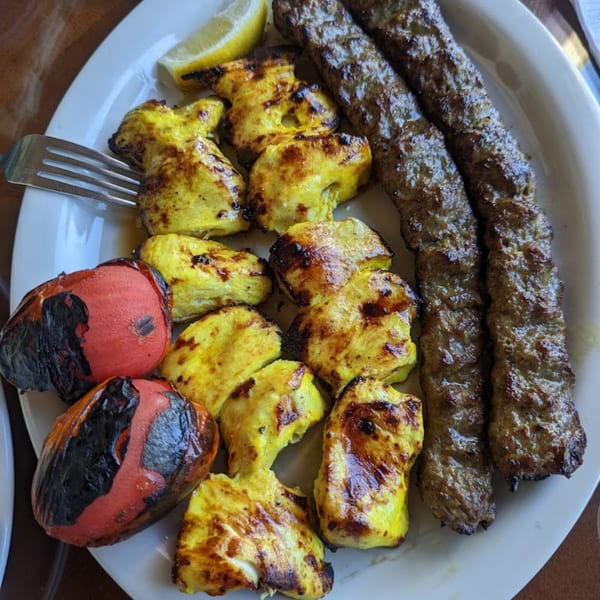 A plate of Persian kabob barg and chicken kabob with grilled tomatoes and a lemon wedge.